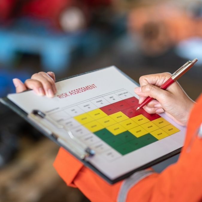Worker in an orange jacket holds a clipboard with a color-coded risk assessment matrix and a red pen in foreground